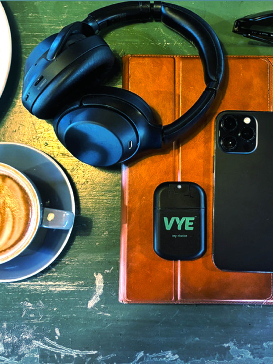 A VYE mist device sits on a café table beside headphones, coffee and a phone.