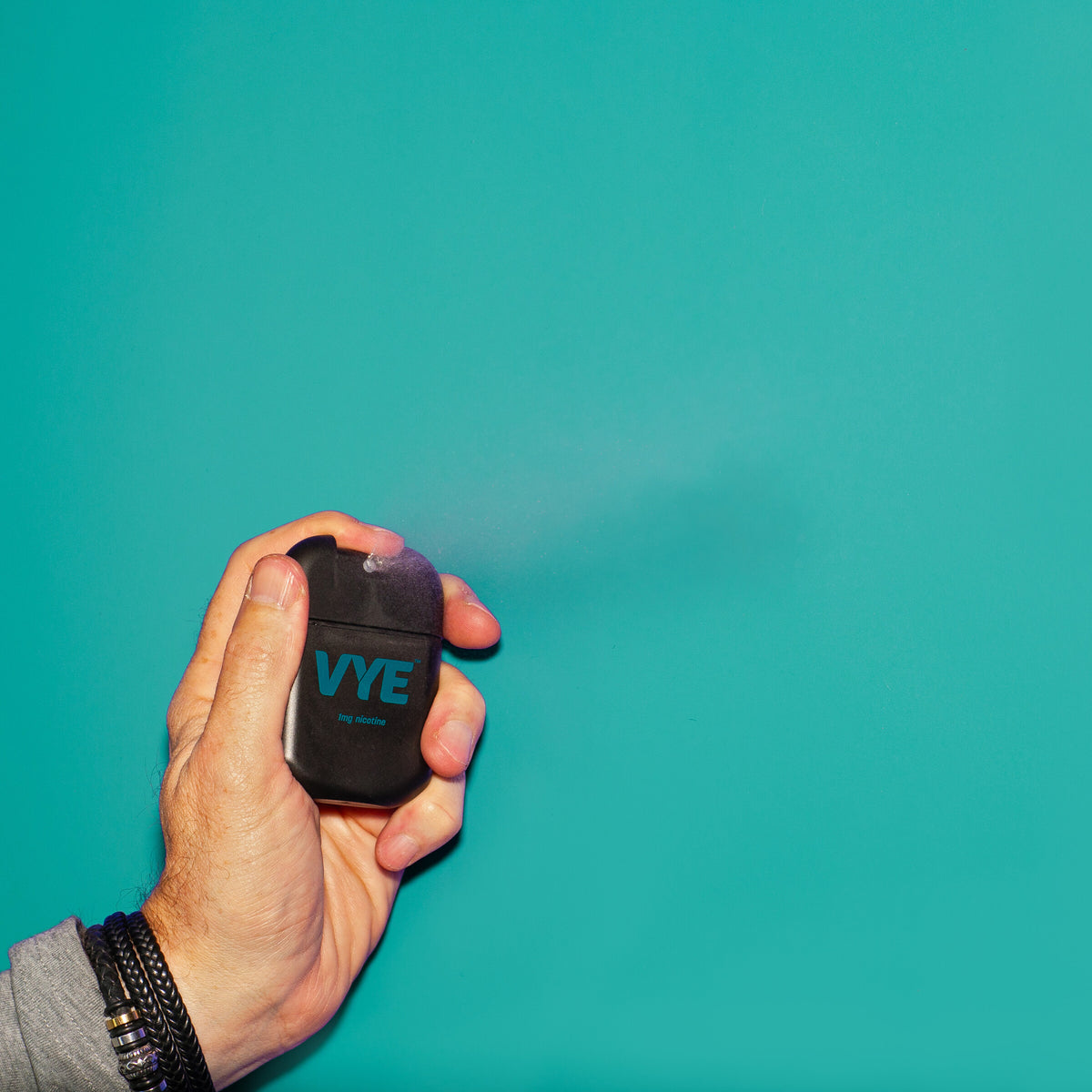 Hand using a black VYE sprayer with mist visible from the nozzle, against a teal background.